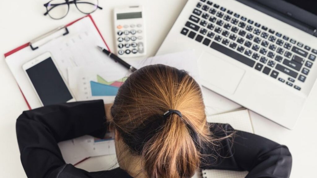 Business Frau am Arbeitsplatz. Sie hat ihren Kopf auf ihrem Schreibtisch abgelegt. Man sieht Laptop, Handy und Arbeitsunterlagen.